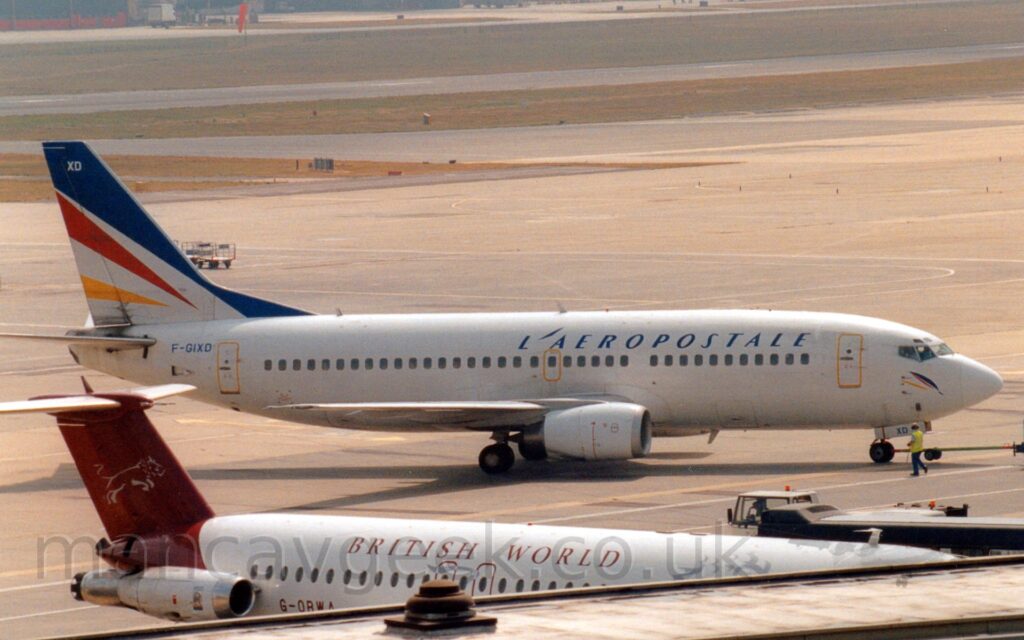 The tail diagonal blue, red, and yellow stripes, outlined in white. The white upper fuselage and purple tail of another plan can be seen parked on the other side of a terminal building in the foreground, while extensive areas of apron in the background give way to grass and a black runway in the distance.