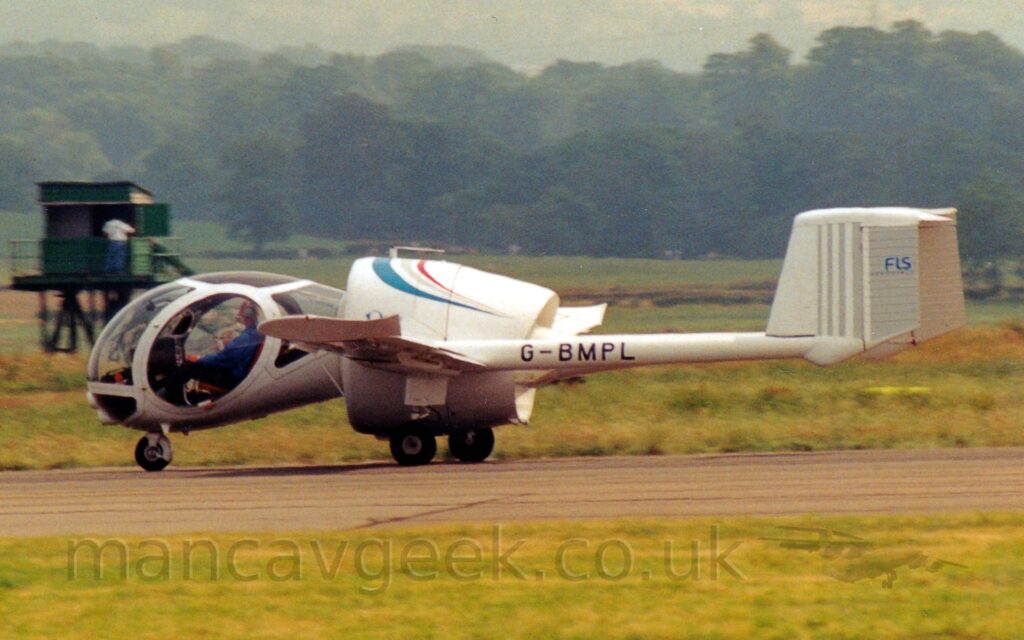 Side view of a single-engined light aircraft taxiing from right to left along a black runway. The plane is mostly white, with a piston engine inside a large shroud in the middle, looking much like a jet engine, with wings sticking out from the side, long twin boom extending from the rear of the wings carrying the tail. The black registration "G-BMPL" is shown on these booms. The cockpit is a complex, heavily glazed ovoid, almost a classic tear-drop shape, sticking out from the front of the engine shroud, and giving the plane an almost insectoid appearance. Lush green grass lines the runway in the foreground, with more grass in the background leading up to trees in the distance, under hazy grey skies.