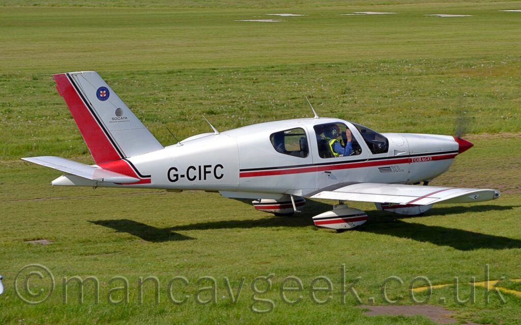 Side view of a single engined light aircraft taxiing from left to right on grass. The plane is mostly white, with a red and black stripe running along the body, turning up into the tail where the red fills the rear edge. A similar stripe runs along the tops of the spats on the undercarriage. Two people can be seen in the cockpit, the person on the left wearing a blue jumper and a yellow hi-viz vest. Green grass fills the background.