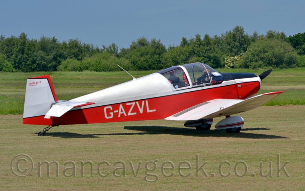 A single engined light aircraft parked facing to the right on grass. The plane has a white top half and red lower half, with a thin black stripe separating the 2 parts, and the white registration "G-AZVL" on the lower rear fuselage. The white wings have the outer panels cranked up slightly, with the leading edges and tips painted red. The tricycle landing gear has white spats with a red stripe over the main wheels. Trees and bushes in the distance mark the airfield perimeter, under very blue skies.