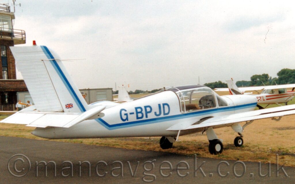 Side view of a white, single engined light aircraft parked on grass facing to the right and away from the camera, although it's rear fuselage is hanging out over a tarmac taxiway. There is a 2-tone blue stripe running along the body, sweeping up along the forward edge of the tail. The registration "G-BPJD" is on the rear fuselage in a dark blue. In the background, a brown brick control tower is on the left of the frame, with other light aircraft parked behind this plane in the middle and on the right. Trees line the airfield perimeter, under flat grey skies.