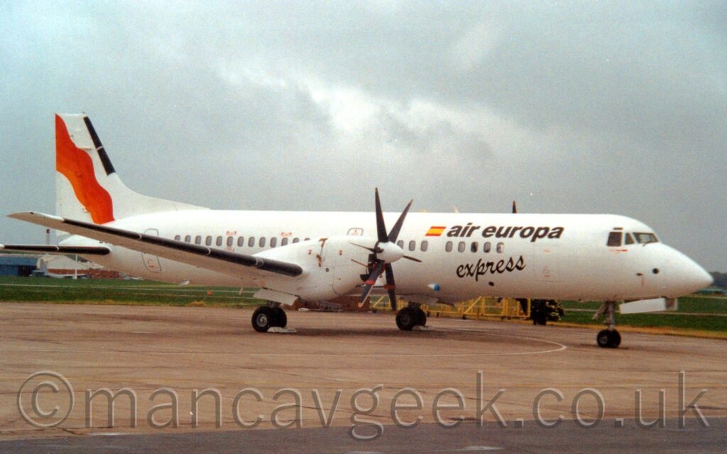 Side view of a twin propellor-engined parked on grey concrete apron facing to the right. The plane is almost entirely white, with black "Air Europa" titles on the upper forward fuselage next to a small Spanish flag, with black, seemingly hand-written "Express" text below. An orange and red wavy diagonal stripe is on the white tail. Grey concrete apron fills the foreground, with grass in the background visible under the plane, with cloudy grey sky above.