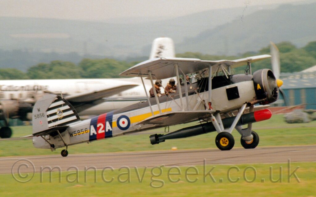 Side view of a large, single engined military torpedo-bomber bi-plane taxiing from left to right along a grey runway. The plane is largely light grey, with dark grey on the top of the body, with a yellow stripe running along the body from under the cockpit, a blue, white and red roundel on the rear fuselage just aft of the cockpit, and a blie, red, and blue diagonal band just behind that, overlaid with the white text "A2A" The black serial "W5856" is under the horizontal stabiliser, with a series of black and white stripes on the overwise grey tail just above it. There is a large black torpedo with a red nose-cone hanging under the fuselage, between the main wheels. The crew of 3, all bundled up in the iconic brown leather flying jackets and leather helmets, sit in open cockpits, the 2 men sitting behind the pilot seeming to hang on for grim death. Green grass fills the foreground, with a white piston-engined airliner in the background on the left, and a gey and blue hangar on the right, and trees and hills in the distance under grey sky.
