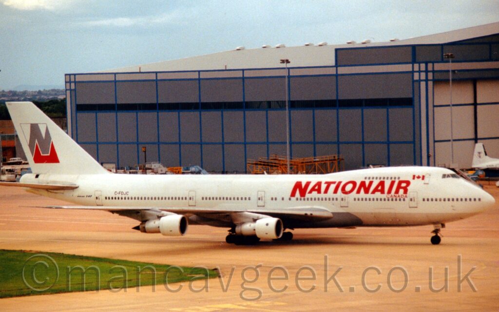 Side view of a very large, white, 4 engined jet airliner taxiing from left to right. The plane has a bare metal belly, and large red "NationAir" titles on the upper forward fuselage. The white tail has the grey outline of a square with a large grey capital letter "N" in the middle, the negative space at the bottom coloured bright red. A large grey hangar fills most of the background from the right, under a hazy grey sky.