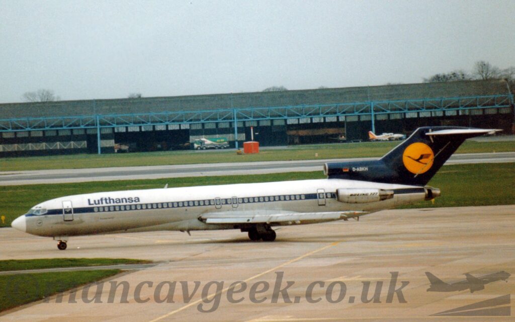 Side view of a 3 engined jet airliner taxiing from right to left. The top of the fuselage is white, as is the nose cone, with a dark blue stripe running along the body, covering the passenger cabin windows, and a bare metal lower body. The centre engine inlet and tail are dark blue, with the white registration "D-ABKH" next to a small German flag, a yellow circle containing a stylised blue flying bird. The other engines, mounted on the sides of the rear fuselage, are in a bare metal finish, with "Boeing 727" titles. Grey concrete apron fills the foreground, with a patch of green grass on the left, with more grass and a runway leading up to a wide, black hangar with blue metal support struts, and a couple of light aircraft parked outside. Flat grey sky fills the rest of the frame.
