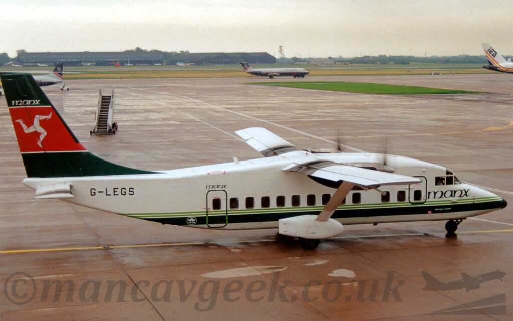 Side view of a rather squat and boxy, high-winged, twin propellor-engined airliner taxiing from left to right. The plane is largely white, with a couple of thin, green pinstripes over a thicker, dark green stripe at the bottom of the fuselage, green "Manx" titles under the cockpit windows and above the main passenger door at the rear, and the black registration "G-LEGS" at the upper rear of the fuselage. The tail is the same dark green, with a thick red stripe outline in white running through the middle, with a white, armoured Triskelion - 3 legs seemingly joined at the hip and appearing to run in a circle - in the centre. Large white sponsons connected to the bottom of the fuselage cover the main wheels, with a white strut coming up and out from the top, supporting the outer panels of the wings. Reddish-grey squares of concrete apron fill the foreground and much of the background, with a strip of grass running through the middle-right, and 3 planes parked in the background. More grass in the distance leads up to several black hangars with cars and planes parked outside, and more than a few trees, under bright but cloudy skies.