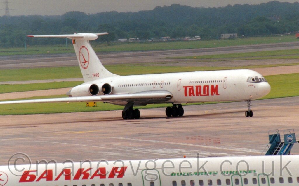 Side view of a 4 engined jet airliner taxiing from left to right along a wide grey taxiway. The plane is mostly white, with red "Tarom" titles on the lower forward fuselage, and the red registration "YR-IRD" at the base of the white tail, as well as the outline of a red circle containing a stylised red flying bird in the middle on the tail. The engines are mounted in horizontal pairs on the sides of the rear fuselage, just below the tail. Grey concrete apron fills the foreground, with the white upper fuselage of another airliner right at the bottom of the frame. Large areas of green grass mixed with taxiways and a black runway fill the background, leading off to trees in the distance, under grey sky.