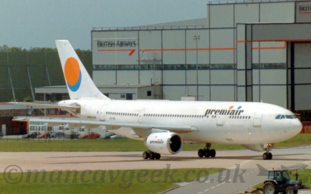 Side view of a white, twin engined jet airliner taxiing from left to right along a grey taxiway. There are black, lower-case "PremiAir" titles on the upper forward fuselage, with orange and blue dots above the letters "i", repeated in a smaller form on the sides of the engine pods under the wings, and a much larger orange dot on the tail, overlaid on a similarly sized blue dot. Geen grass fills the foreground with a grey roadway cutting through on the right. A large grey hangar with the British Airways logo on the side dominates the background on the right, with a smaller brown and grey building on the left, with a boxy, green metallic frame behind that, with grey-blue sky filling the rest of the frame.