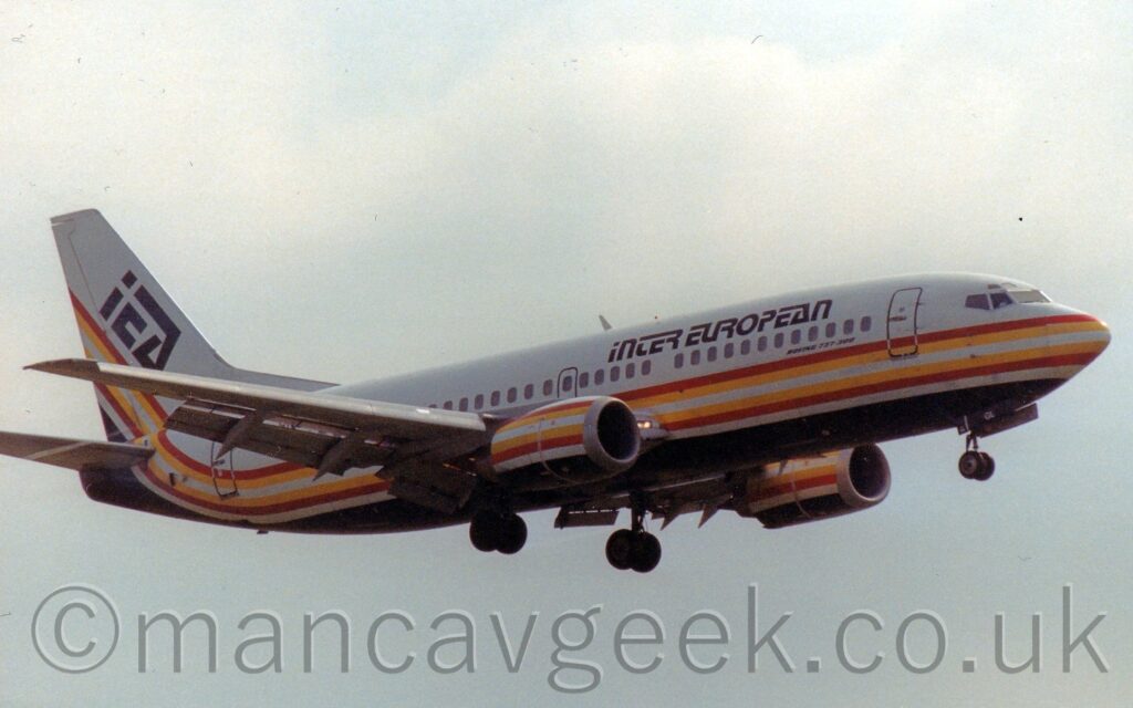 Twin engined jet airliner flying from left to right at a very low altitude, with undercarriage lowered and flaps extended from the rear of the wings, suggesting it is about to land. The plane is mostly white, with a red-yellow-white-yellow-red stripe running along the body under the passenger windows, and a black belly, the stripe and belly sweeping up into the lower reaches of the tail, and repeated on the sides of the engines under the wings. There are blocky, black, "Inter European" titles on the upper forward fuselage, and large, black, diagonal "IEA" titles on the tail. Flat grey sky fills the rest of the frame.