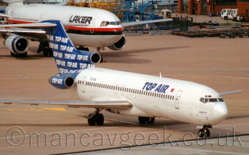 Front view of a 3 engined jet airliner with the engines mounted on the sides of the rear fuselage, taxiing from left to right. The plane is mostly white, with blue "Top Air" titles on the upper forward fuselage, next to a small red and white Turkish flag, and the black registration "TC-IYA" on the upper rear. The tail and rear fuselage are the same blue, with the words "Top Air" repeated in staggered lines, as well as on the sides of the engines mounted on the side of the rear fuselage. The grey roof of a terminal building stretches across the bottom right of the foreground, with large areas of grey concrete apron filling the rest of the foreground and most of the background, with a larger, white, 3 engined jet airliner with black "Laker" titles parked in the top left.
