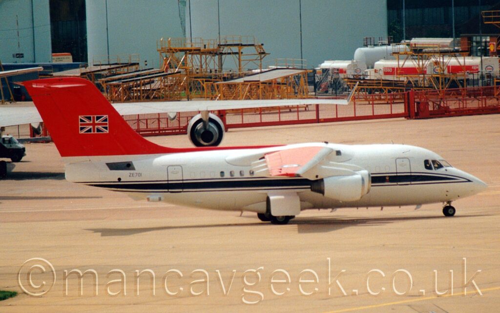 Side view of a high-winged, 4 engined jet airliner taxiing from left to right. There is a dark blue stripe running along the body, under the passenger cabin witndows, and the black military serial "ZE701" on the upper rear fuselage. The tail is red, with a UK flag outlined in white bang in the centre. Grey concrete apron fills the foreground, with the wing of a very large jet airliner stretching across the frame in the background. Duck egg blue fuel tanks are right at the back of the frame, with a collection of yellow metal frames parked in front, behind a red mesh metal fence. Some red and white fuel trucks are parked behind the red fence on the right of the frame.