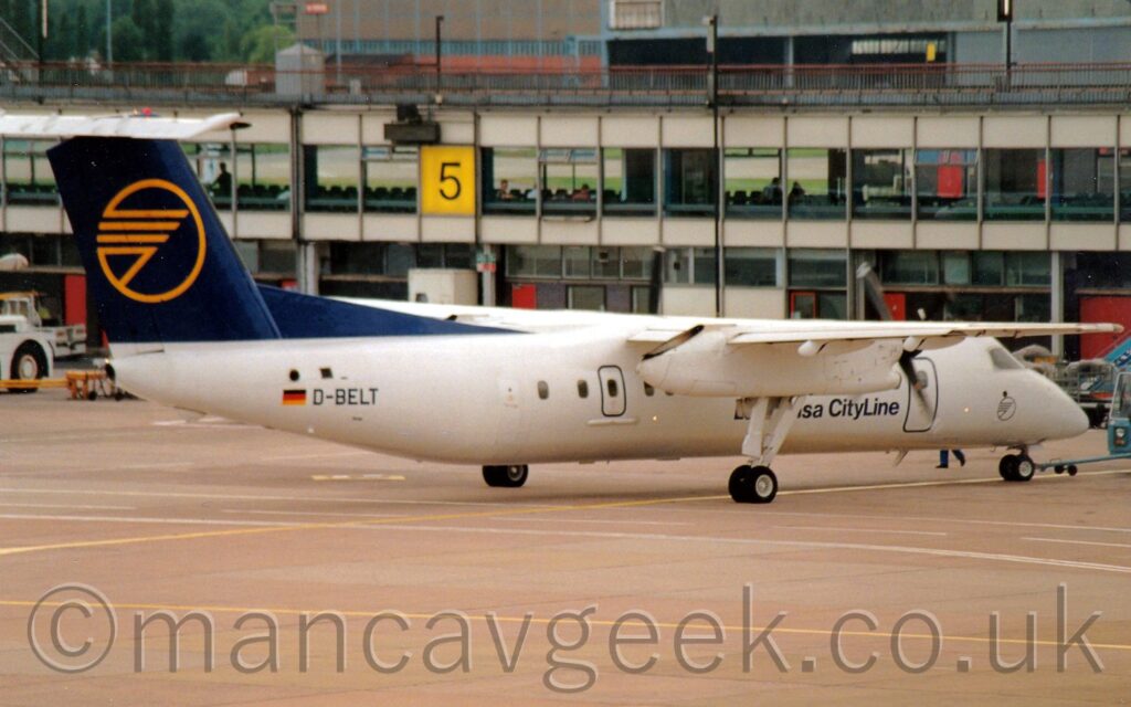 Rear side view of a high-winged, twin propellor-engined airliner, facing to the right but being pushed back to the left by a low-slung blue tug peeking into the frame on the right. The plane is largely white, with partially obscured titles "sa Cityline" on the lower forward fuselage, and the black registration "D-BELT" on the rear fuselage, just in front of a small German flag. The tail and the fillet just in front of it are dark blue, with the yellow outline of a circle containing a highly stylised yellow bird mostly made up of straight lines. Grey concrete apron fills the foreground, with a heavily glazed terminal building stretching all the way across the frame, with one of the windows replaced with a large yellow square with the black number "5" in the middle. A grey hangar with a wide open door fills most of the top of the frame from the right, with trees on the left.