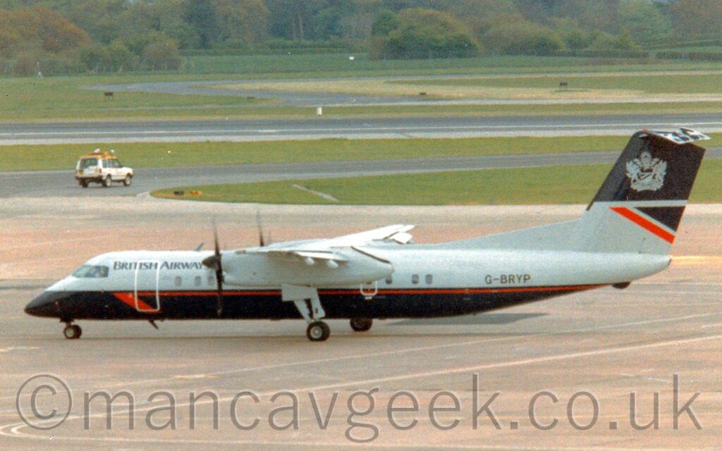 Side view of a high-winged, twin propellor-engined airliner taxiing from right to left across a large expanse of grey concrete apron. The plane's upper half is grey, with black "British Airways" titles on the upper forward fuselage, and the black registration "G-BRYP" on the upper rear. The lower half is dark blue, almost black, with a thin red stripe running along the body near the grey join, ending in a red half arrowhead just aft of the cockpit windows. The engine pods under the wings are also grey. The bottom of the tail is grey, with a red and dark blue triangle on the trailing edge, while the top half is dark blue, with a grey Heraldic crest in the middle. Grey apron fills the foreground at the bottom of the frame, with a roadway marked on it for ground vehicles, with a large grassed area in the background split by taxiways and a runway. A white car with a yellow roof and orange lights mounted above the roof is driving along one of the taxiways on the left of the background. More grass on the other side of the runway leads up to trees in the distance.