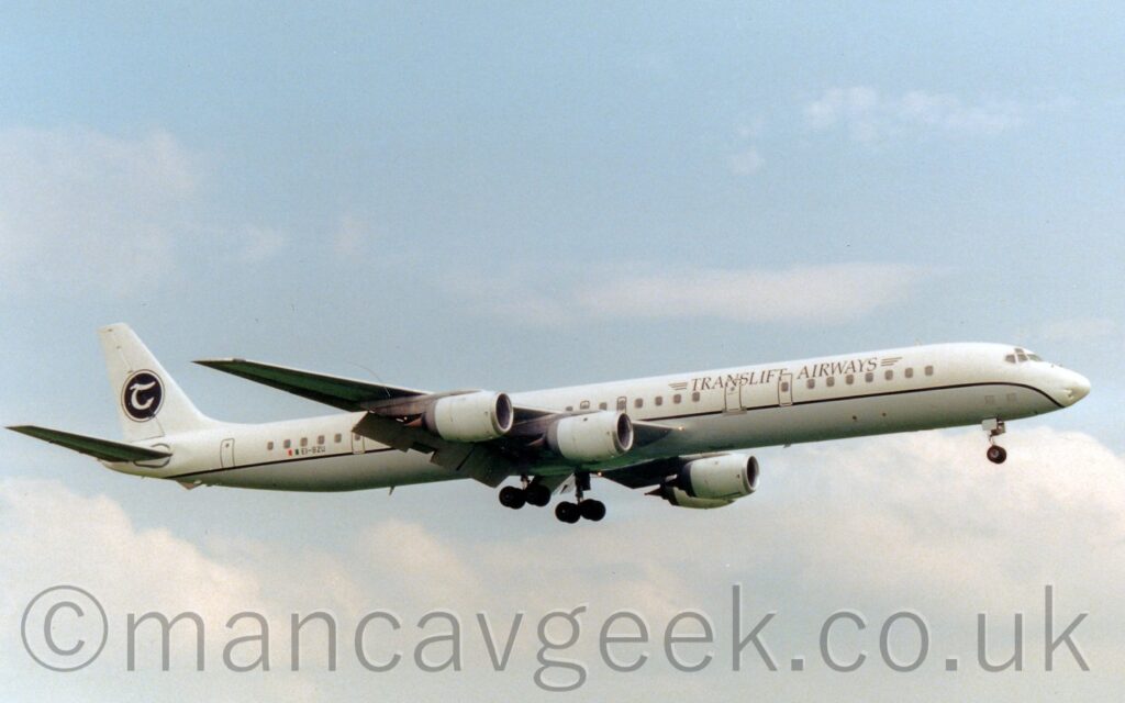A long and thin, 4 engined jet airliner flying from left to right at a low altitude, with undercarriage lowered, and large flaps hanging down from the rear of the wings, suggesting it is about to land. The engines look massive on this plane, almost comically so. The plane is largely white, with a grey-blue belly, and a thin black stripe running along the body separating the 2 sections. There are black "TransLift Airways" titles on the upper forward fuselage, and a the black registration "EI-BZU" on the lower rear, next to a small Irish flag. A black circle, outlined in black, and containing a stylised white letter "t" sits in the middle of the white tail. Fluffs of white cloud with hints of blue peeking through fill the rest of the frame.