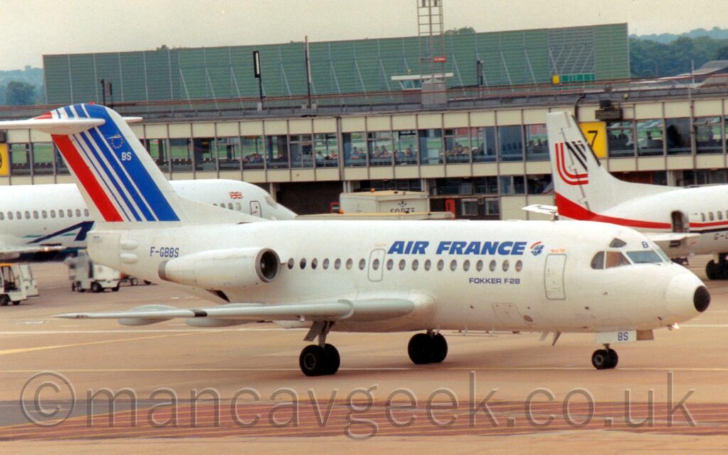 Side view of a twin engined jet airliner with the engines mounted on the sides of the rear fuselage, taxiing from left to right. The plane is almost entirely white, with blue "Air France" titles on the upper forward fuselage, next to a small image of a red and blue sea horse. The blue registration "F-GBBS" is on the side of the rear fuselage, just behind the engines. Blue concrete apron fills the foreground, with a couple of airliners parked nose-in against a terminal building that stretches across the frame in the background. A boxy green metal structure looms over everything in the background, under hazy grey sky.