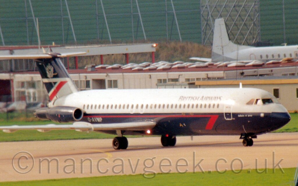 Side view of a twin engined jet airliner with the engines mounted on the sides of the rear fuselage, taxiing from left to right along a grey taxiway. The plane has a grey upper half and dark blue lower half, with a thin red stripe running along the top of the blue section, ending in a red half-arrowhead at the front, just aft of the forward cabin door. There are black "British Airways" titles on the upper forward fuselage, and the grey text "Bailiwick of Jersey" on the lower forward fuselage, just below the rearmost cockpit window. The grey registration "G-0AVMP" is on the lower rear fuselage, just below and in front of the dark blue engines on that side of the body. The rather squat tail is grey at the bottom, with a dark blue and red triangle at the rear, while the top is the same dark blue, with a grey heraldic crest in the middle. Green grass lines the side of the taxiway in the foreground, with more in the background, giving way to a grey single-storey grey building with a flat red roof and raised skylights,, with a white plane parked, facing out of frame, beyond that on the right. A large, boxy, green metal frame looms over everything else in the background.