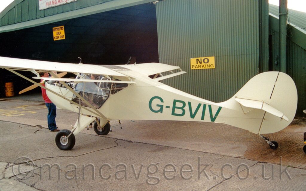 Rear side view of a creamy green, high-winged, single-engined light aircraft parked facing to the left on grey tarmac. The large green registration "G-BVIV" is on the side of the rear fuselage. A large green hangar fills the background, it's wide, open entrance on the left of the frame.
