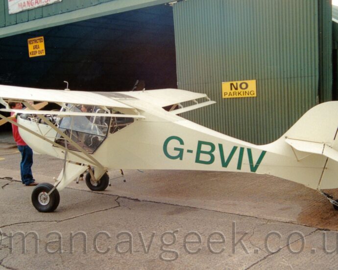 Rear side view of a creamy green, high-winged, single-engined light aircraft parked facing to the left on grey tarmac. The large green registration "G-BVIV" is on the side of the rear fuselage. A large green hangar fills the background, it's wide, open entrance on the left of the frame.