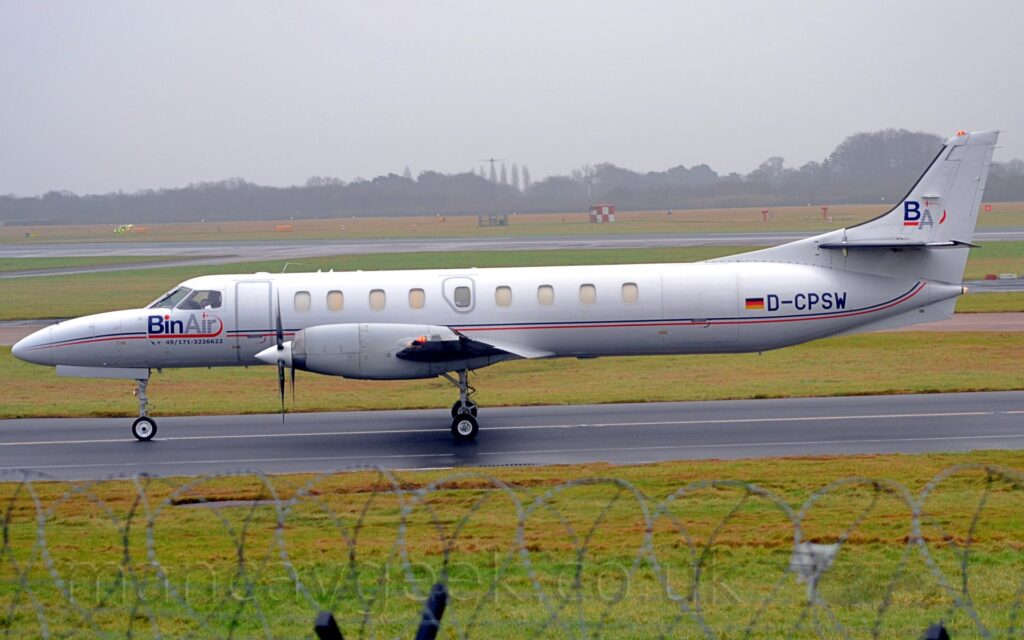 Side view of a long and thin twin propellor-engined airliner taxiing from right to left along a black taxiway. The plane is mostly white, with a thin red and blue stripe running along the body, and large blue "BinAir" titles under the cockpit windows. There are large blue letters "BA" on the tail, and the black registration "D-CPSW" on the rear fuselage, next to a large German flag. Green grass lines the taxiway in the foreground, with a roll of razorwire on top of a fence visible right at the bottom. More grass lines taxiways and a runway in the background, leading up to trees vanishing in to mist in the distance.