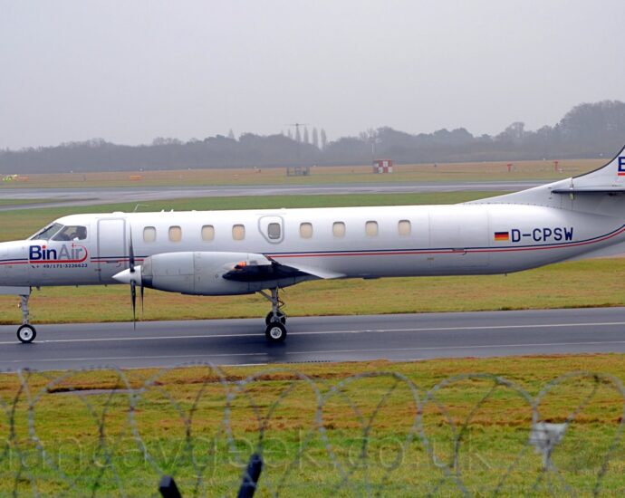 Side view of a long and thin twin propellor-engined airliner taxiing from right to left along a black taxiway. The plane is mostly white, with a thin red and blue stripe running along the body, and large blue "BinAir" titles under the cockpit windows. There are large blue letters "BA" on the tail, and the black registration "D-CPSW" on the rear fuselage, next to a large German flag. Green grass lines the taxiway in the foreground, with a roll of razorwire on top of a fence visible right at the bottom. More grass lines taxiways and a runway in the background, leading up to trees vanishing in to mist in the distance.