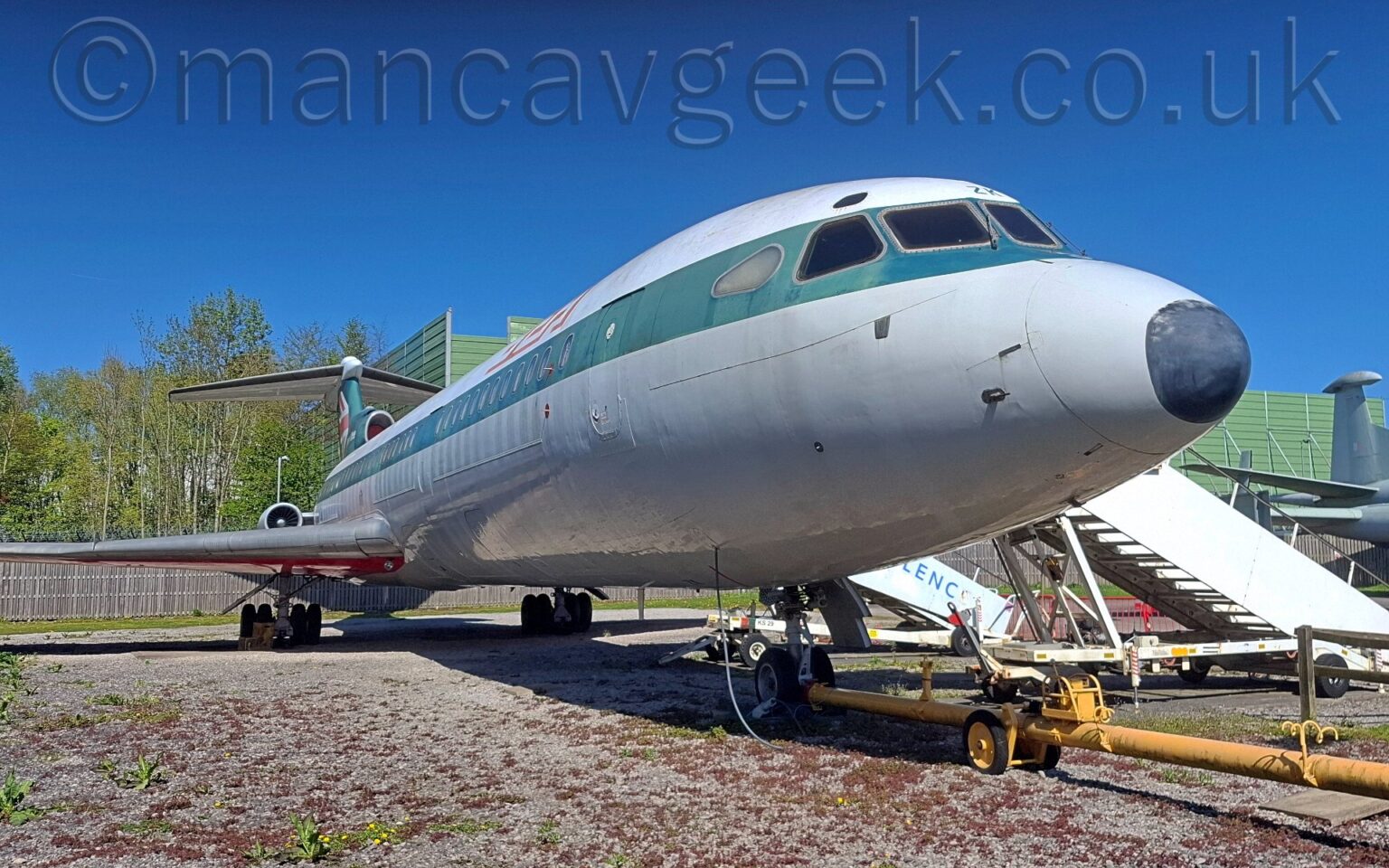 Low front view of a 3 engined jet airliner parked facing to the right on gravel. The plane is white on top, with a grey lower half, and a thick stripe that used to be dark blue but is now faded to dark green running along the body, covering the passenger cabin windows. There are red and white letters "BEA" on the upper forward fuselage, and a partial UK flag on the now-green tail. The underside of the wings is red. A long yellow pushback pole is attached to the nosewheel. White airstairs are attached to the front and rear cabin doors on the far side of the plane. Trees fill the background on the left of the frame, with a rather square and boxy green metal frame taking up most of the rest of the background, and the grey tail of another plane on the right, all under gorgeous blue sky.