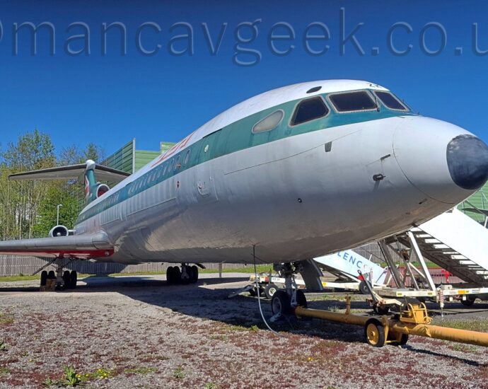 Low front view of a 3 engined jet airliner parked facing to the right on gravel. The plane is white on top, with a grey lower half, and a thick stripe that used to be dark blue but is now faded to dark green running along the body, covering the passenger cabin windows. There are red and white letters "BEA" on the upper forward fuselage, and a partial UK flag on the now-green tail. The underside of the wings is red. A long yellow pushback pole is attached to the nosewheel. White airstairs are attached to the front and rear cabin doors on the far side of the plane. Trees fill the background on the left of the frame, with a rather square and boxy green metal frame taking up most of the rest of the background, and the grey tail of another plane on the right, all under gorgeous blue sky.