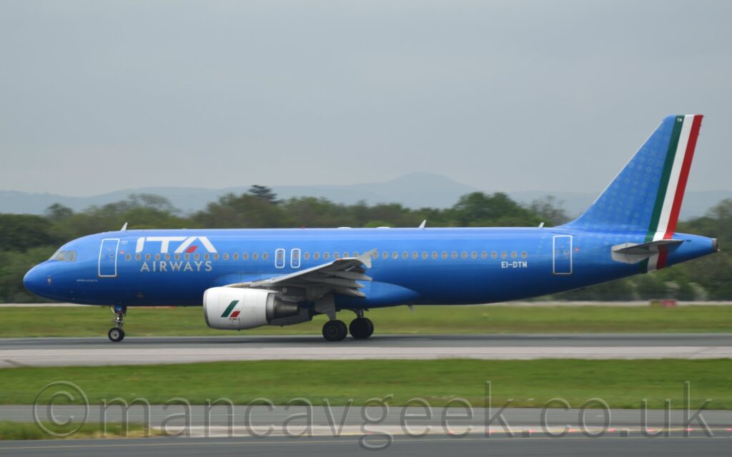 Side view of a very blue, twin engined jet airliner acelerating along a black runway during it's take-off roll. The plane is in a metallic blue livery, with large white "ITA" titles on the upper forward fuselage, over small white text "Airways", amd the white registration "EI-DTM" on the lower rear fuselage. The tail is the same blue, with green, white, and red bands representing the Italian flag running down the trailing edge and round the tail-cone. Green grass and grey taxiways fill the foreground, with trees in the distance, under grey sky.
