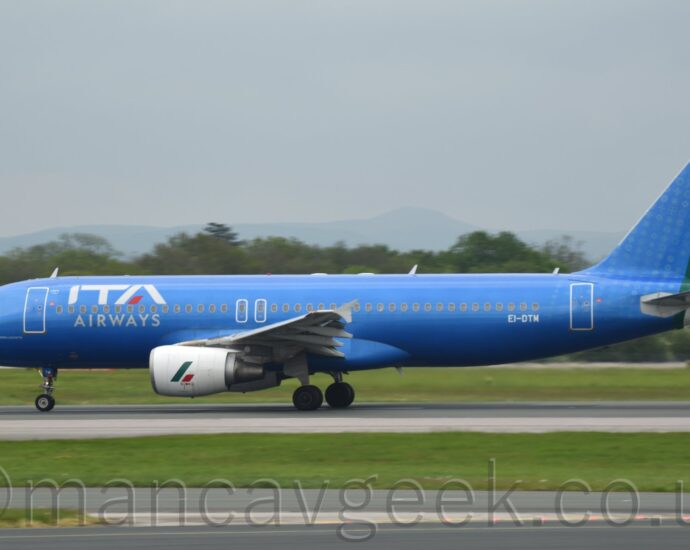 Side view of a very blue, twin engined jet airliner acelerating along a black runway during it's take-off roll. The plane is in a metallic blue livery, with large white "ITA" titles on the upper forward fuselage, over small white text "Airways", amd the white registration "EI-DTM" on the lower rear fuselage. The tail is the same blue, with green, white, and red bands representing the Italian flag running down the trailing edge and round the tail-cone. Green grass and grey taxiways fill the foreground, with trees in the distance, under grey sky.