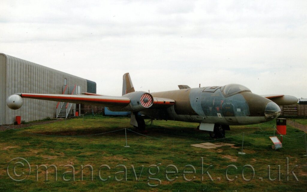 Front 3/4 view of a twin engined military jet bomber parked facing to the right on grass, on display outside a museum. The plane is covered in a grey and brown camouflage scheme, with a pale blue-green belly, the same colour being used on the undersides of the engine pods buried in the mid-points of the wings, under the wings themselves, and all over the external fuel tanks mounted under the wing-tips. The leading edges of the wings are painted bright orange, and the are red and white striped blanking plates inside the engine inlets. The planes nose is a glass bubble, and a large bubble canopy covers the cockpit. Green grass fills the foreground, with a thin string of rope suspended from several poles on the ground marking an inaccessible area around the plane and a red and brown rubbish bin under the planes left wing. A grey, corrugated metal building fills the background on the left, with bright but hazy sky filling the rest of the frame.