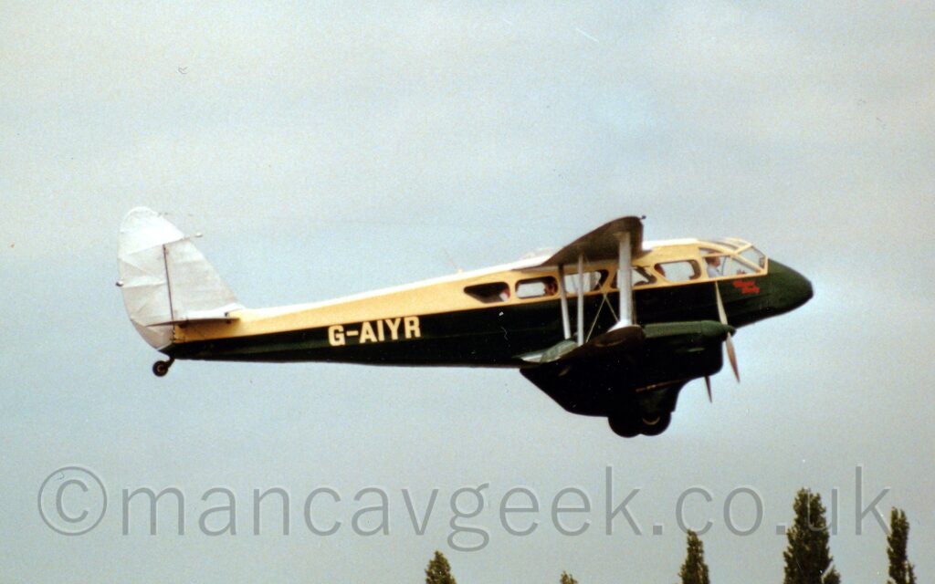 Side view of a twin propellor-engined, bi-plane airliner of pre-World War 2-era vintage flying from left to right at a low altitude, doing one of a series of flybys. The plane is a creamy yellow on top, with a green lower half and engines mounted on the lower wings. The yellow registration "G-AIYR" is on the side of the rear fuselage. The tops of some trees in the distance can be seen at the bottom of the frame, with cloudy grey sky with hints of blue threatening to peek through filling the rest of the frame.