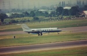 Slightly blurry and grainy, side view of a twin engined BizJet taxiing from left to right. The plane is mostly white, with a dark blue belly, and a yellow stripe running along the body, with another yellow stripe on the tail. A black runway bordered by large areas of grass fills the foreground, with more grass leading up to trees in the background.