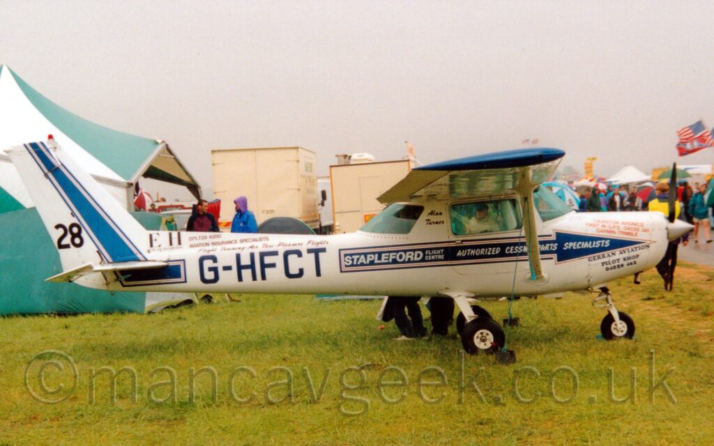 Side view of a high-wing, single engined light aircraft parked facing to the right on grass. There are stickers, mostly with blue text, advertising several companies that most people would have never heard of on the sides of the plane, along with a blue stripe running along the body and up into the tail, the black registration "G-HFCT" on the side of the rear fuselage, and the black number "28" right at the lower rear of the tail. Green grass fills the foreground, with a large green tent on the right of the background, and a couple of cream trucks in the middle, under slightly grim grey sky.