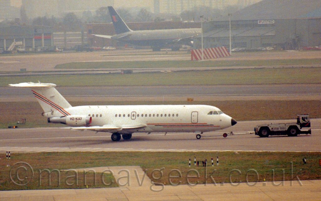 Side view of a white, twin-engined jet airliner with the engines mounted on the sides of the rear fuselage, being towed from left to right along a grey taxiway by a dark blue and grey tug attached to the nosewheel by a long white pole. The plane has a thin, red, black, and red stripe running along the body before sweeping up into the tail. The black registration "HZ-KA7" is on the side of the white engine pod on the rear fuselage. Taxiways and large grassed areas fill the foreground, with a grey jet airliner trying to hide behind a hangar in the distance, on the other side of a black runway and more grass.