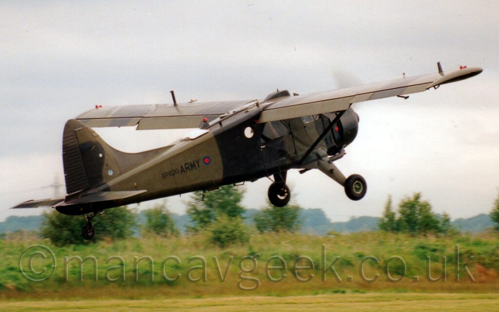 Side view of a high-winged, single propellor-engined military transport plane flying from left to right at a very low altitude, just metres above a runway, with flaps deployed from behind the wing, and the nose raised sharply, suggesting it has just taken off. The plane is covered in a dark and light green camouflage pattern, with the serial "XP820" next to black "Army" titles, itself next to a small red and blue roundel, on the rear fuselage. Bushes and trees in the background mark the airfield perimeter, with more trees fading in to hazy grey skies in the distance.
