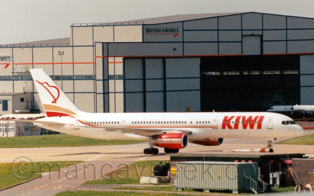 Side view of a white, twin engined jet airliner taxiing from left to right. The plane has thin gold and red stripes running along the body with large red "Kiwi" titles on the forward fuselage, and a the red outline of a stylised kiwi bird. The engine pods under the wings are red, with white and gold stripes. Green grass fills the left of the foreground, with some low buildings and a grey shipping container on the right. A large grey hangar with blue and red piping and blue British Airways titles dominates the background, with a blue and grey plane parked outside it on the right, and another plane in similar colours inside the open doors. Grey sky with hints of blue fills the rest of the frame.