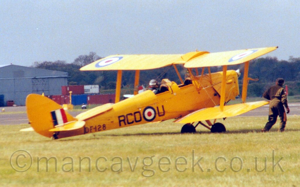 Side view of a bright yellow, single-engined biplane taxiing from left to right on grass. The plane has a large, blue, white, and red roundel just below and aft of the rear cockpit, with the black letters "RCO" on one side, and "U" on the other. Similar roundels are on the outer panels of the top of the upper wing. The black serial "DF128" is on the lower rear fuselage, under the tail. The tail is also bright yellow, with a large, square fin flash in blue, white, and red. The pilot is sitting in the open rear cockpit, and a man wearing green fatigues is walking alongside the right wingtips. Green grass fills the foreground, with a grey hangar and red and blue shipping containers in the background on the left, and trees on the right, under flat grey sky.