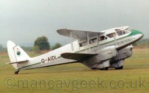 Side view of a twin propellor-engined, bi-plane, pre-World War-2 era airliner taxiing from left to right along a grass taxiway. The plane is largely white, with a grey lower half, and a green and black stripe running along the body that widens and sweeps up over the nose at the front. The black registration "G-AIDL" is on the side of the rear fuselage. There are green "Caernarfon Air World" titles on the lower forward fuselage. Several passengers can be seen looking out and waving from the rounded triangular cabin windows. Green grass fills the foreground and a large part of the background, with trees and bushes in the distance, under flat grey sky.