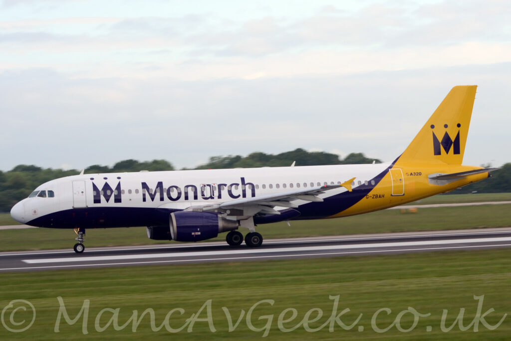 Side view of a twin engined jet airliner moving from right to left at an accelerating pace. The plane is mostly white, with a purple belly and engine pods under the wings, and bright yellow rear fuselage and tail. There is a stylised purple letter "M" on the tail, designed to resemble a crown, which is repeated in a larger form on the tail. Over the wings, there are large purple "Monarch" titles, and the black registration "G-ZBAH" on the yellow upper rear fuselage, under the tail. Green grass fills the foreground, with more on the far side of the runway, with trees in the distance under pale blue sky with plenty of white and grey fluff.