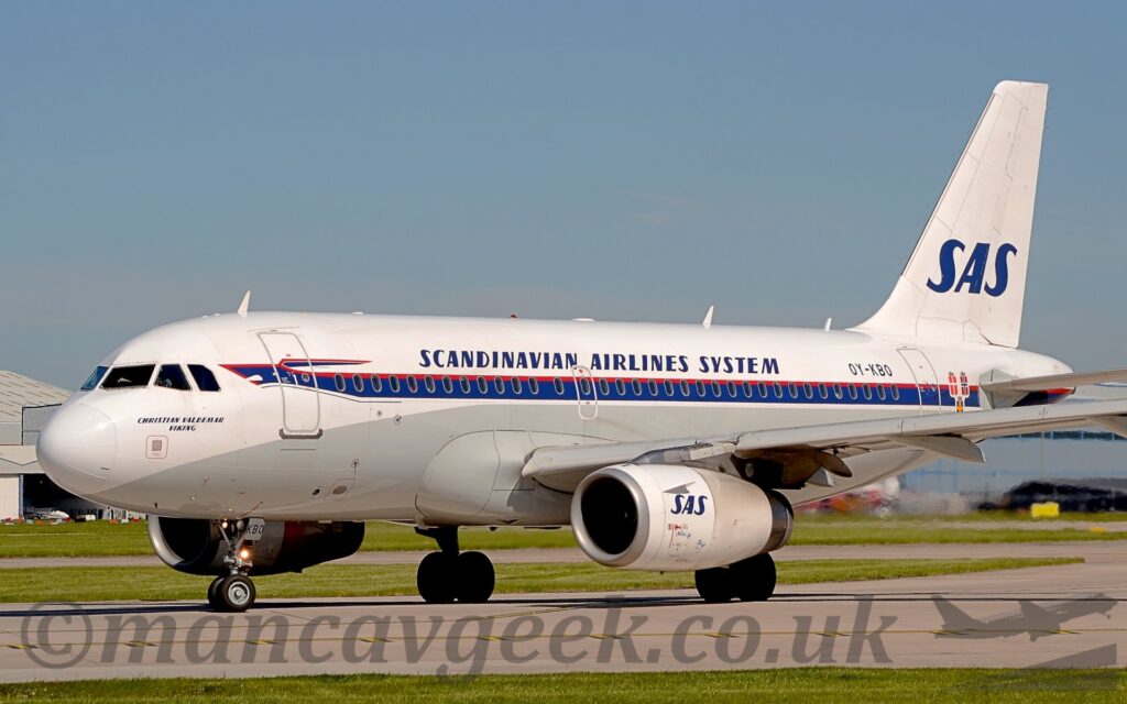 Front side view of a twin engined jet airliner taxiing from right to left, along a grey runway. The plane is mostly white, with a grey lower half, and a dark blue stipe topped by a thinner red stripe, resembling a Viking long boat at the front, that covers the passenger cabin windows. The blue name "Christian Valdemar Viking" is on the side of the nose, under the cockpit windows. There are dark blue "Scandinavian Airlines System" on the upper fuselage, over the wings, and the black registration "OY-KBO" on the upper rear, above the rearmost windows. There are large, dark blue letters "SAS" on the white tail, repeated in a smaller form on the sides of the white engine pods, mounted under the wings. Grass lines the sides of the runway at the bottom of the frame, with blue sky filling the top.