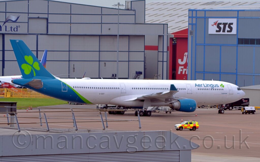 Side view of a twin engined jet airliner parked facing to the right on a large section of grey concrete apron. The plane is largely white, with bright green "Aer Lingus" titles on the side of the upper forward fuselage, next to an image of a small green shamrock. A larger version of the shamrock logo appears on the blue-green tail, which also covers the rear fuselage, separated from the white by a bright green stripe that runs diagonally down the rear tail. The green registration "G-EIDY" is on the lower rear fuselage, just aft of the wing. Grey concrete apron fills a large chunk of the foreground, with the roof of a grey Airport building in the bottom left corner. A pair of large grey hangars, one on each side, dominate the background, with the red doors of a third visible in the gap in the middle.