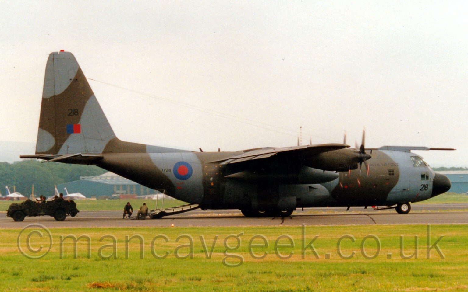 Side view of a high-winged, 34 propellor-engined military transport aircraft parked facing to the right on a black runway, with it's cargo ramp in the rear fuselage deployed, with a vehicle moving along the runway that has obviously just come out of the back of the plane. The plane is covered in a grey and green-brown camouflage scheme, with small "Royal Air Force" titles on the side of the forward fuselage, the large black number "218" under the cockpit windows on the nose. There is a large blue and red roundel on the side of the rear fuselage, followed by the small serial "XV218". The black number "218" is repeated on the tail, over a blue and red fin flash. Green grass fills the foreground, with more grass on the far side of the runway. More grass in the background leads up to a pair of white and blue jet fighters on the left, and blue hangars on left and right, with trees beyond, under flat white sky.