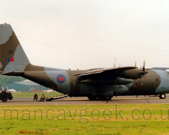 Side view of a high-winged, 34 propellor-engined military transport aircraft parked facing to the right on a black runway, with it's cargo ramp in the rear fuselage deployed, with a vehicle moving along the runway that has obviously just come out of the back of the plane. The plane is covered in a grey and green-brown camouflage scheme, with small "Royal Air Force" titles on the side of the forward fuselage, the large black number "218" under the cockpit windows on the nose. There is a large blue and red roundel on the side of the rear fuselage, followed by the small serial "XV218". The black number "218" is repeated on the tail, over a blue and red fin flash. Green grass fills the foreground, with more grass on the far side of the runway. More grass in the background leads up to a pair of white and blue jet fighters on the left, and blue hangars on left and right, with trees beyond, under flat white sky.