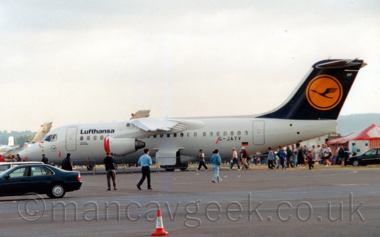 Side view of a high-winged, 4 engined jet airliner on static display at an airshow, parked facing to the left. The plane is almost entirely white, with black "Lufthansa" titles on the upper forward fuselage, and the black registration "G-JAYV" on the lower rear. Below the registration is a panel with a slightly different shade of white, covering the registration it will have after delivery, next to a small German flag. The tail is a dark blue, with a yellow circle in the middle, containing a stylised blue flying bird, and the white letters "RF" in the top right corner. A large expanse of tarmac fills the foreground, with a row of cars on the left, and people walking around in front of the plane. Other planes can be seen in the background on the left of the planes, with a red tent on the right, and crowds of people walking around on both sides. Flat white sky fills the rest of the frame.