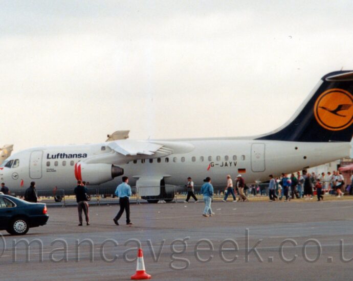 Side view of a high-winged, 4 engined jet airliner on static display at an airshow, parked facing to the left. The plane is almost entirely white, with black "Lufthansa" titles on the upper forward fuselage, and the black registration "G-JAYV" on the lower rear. Below the registration is a panel with a slightly different shade of white, covering the registration it will have after delivery, next to a small German flag. The tail is a dark blue, with a yellow circle in the middle, containing a stylised blue flying bird, and the white letters "RF" in the top right corner. A large expanse of tarmac fills the foreground, with a row of cars on the left, and people walking around in front of the plane. Other planes can be seen in the background on the left of the planes, with a red tent on the right, and crowds of people walking around on both sides. Flat white sky fills the rest of the frame.