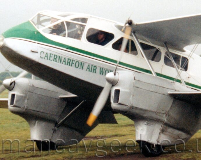 Close up of the forward fuselage of a white, twin engined bi-plane airliner taxiing from right to left on grass. The plane has a grey belly and engines, with a green stripe running along the body and green "Caernarfon Air World" titles on the lower forward fuselage. The pilot is visible in the cockpit, with passengers sitting in the cabin, visible through the triangular windows. Grey sky fills the rest of the frame.