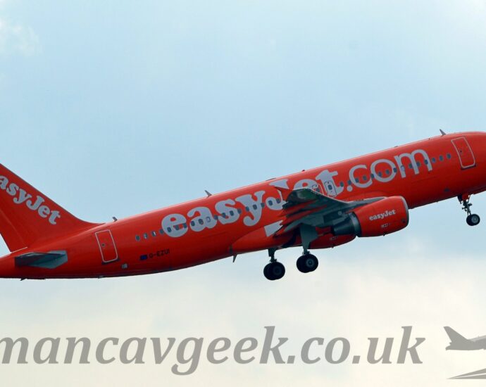 Side view of a bright orange, twin engined jet airliner flying from left to right at a low altitude, with undercarriage still lowered, flaps slightly extended from the rear of the wings, and the nose raised sharply, suggesting it has just taken off. There are large, white "EasyJet.com" titles written across the fuselage, with smaller "EasyJet" titles on the engine pods under the wings, as well as diagonally on the tail. There is a large white number "200" on the forward fuselage just under the cockpit windows, and the black registration "G-EZUI" on the lower rear fuselage. Pale blue sky with white patches of cloud fill the rest of the frame.