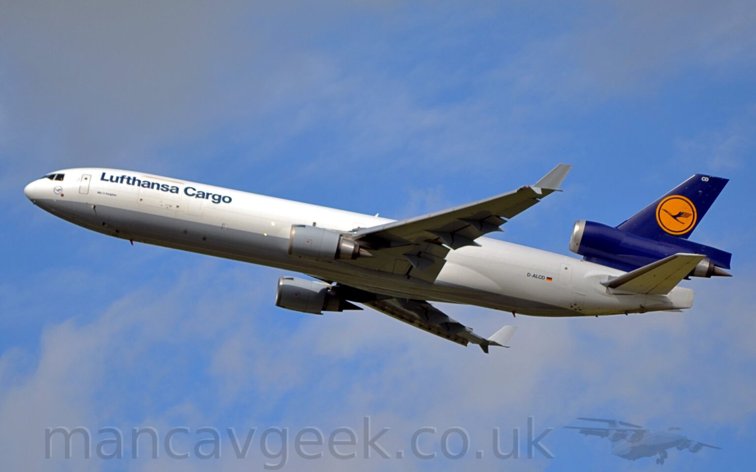 Side view of a 3 engined jet airliner flying from right to left at a low altitude, with undercarriage already tucked away, flaps extended from the rear of the wings, and the nose raised significantly, suggesting that it has just taken off. The plane is mostly white, with a grey belly, and dark blue "Lufthansa Cargo" titles on the upper forward fuselage, and the black registration "D-ALCD" next to a small German flag on the lower rear. The tail and centre engine pod are dark blue, with a yellow circle containing a stylised blue flying bird in the middle. The engine pods mounted under the wings are grey, as are the up-turned wing-tips. Pale blue sky with grey fluffs of cloud fill the rest of the frame.