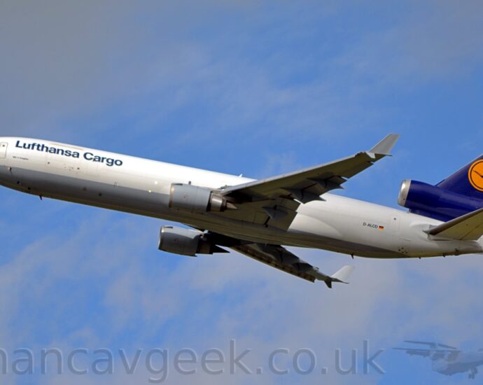 Side view of a 3 engined jet airliner flying from right to left at a low altitude, with undercarriage already tucked away, flaps extended from the rear of the wings, and the nose raised significantly, suggesting that it has just taken off. The plane is mostly white, with a grey belly, and dark blue "Lufthansa Cargo" titles on the upper forward fuselage, and the black registration "D-ALCD" next to a small German flag on the lower rear. The tail and centre engine pod are dark blue, with a yellow circle containing a stylised blue flying bird in the middle. The engine pods mounted under the wings are grey, as are the up-turned wing-tips. Pale blue sky with grey fluffs of cloud fill the rest of the frame.