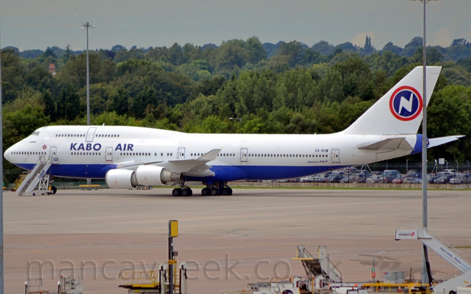 Side view of a very large, 4 engined jet airliner parked facing to the left. The plane is mostly white, with a blue belly, and blue "Kabo Air" titles on the upper forward fuselage, between the upper and lower deck cabin windows. A blue circle with a red outline is on the tail, with a stylised white lightning bolt (or possibly upper case letter "N") in the middle. A tall, grey lighting pole in the foreground partially obscures the tail-cone and tail. Grey concrete apron fills the foreground, with various white airport trucks and mobile airstairs gathered in the bottom right corner. Trees fill the background, under grey skies.