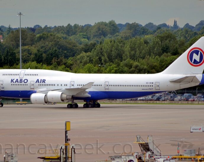 Side view of a very large, 4 engined jet airliner parked facing to the left. The plane is mostly white, with a blue belly, and blue "Kabo Air" titles on the upper forward fuselage, between the upper and lower deck cabin windows. A blue circle with a red outline is on the tail, with a stylised white lightning bolt (or possibly upper case letter "N") in the middle. A tall, grey lighting pole in the foreground partially obscures the tail-cone and tail. Grey concrete apron fills the foreground, with various white airport trucks and mobile airstairs gathered in the bottom right corner. Trees fill the background, under grey skies.